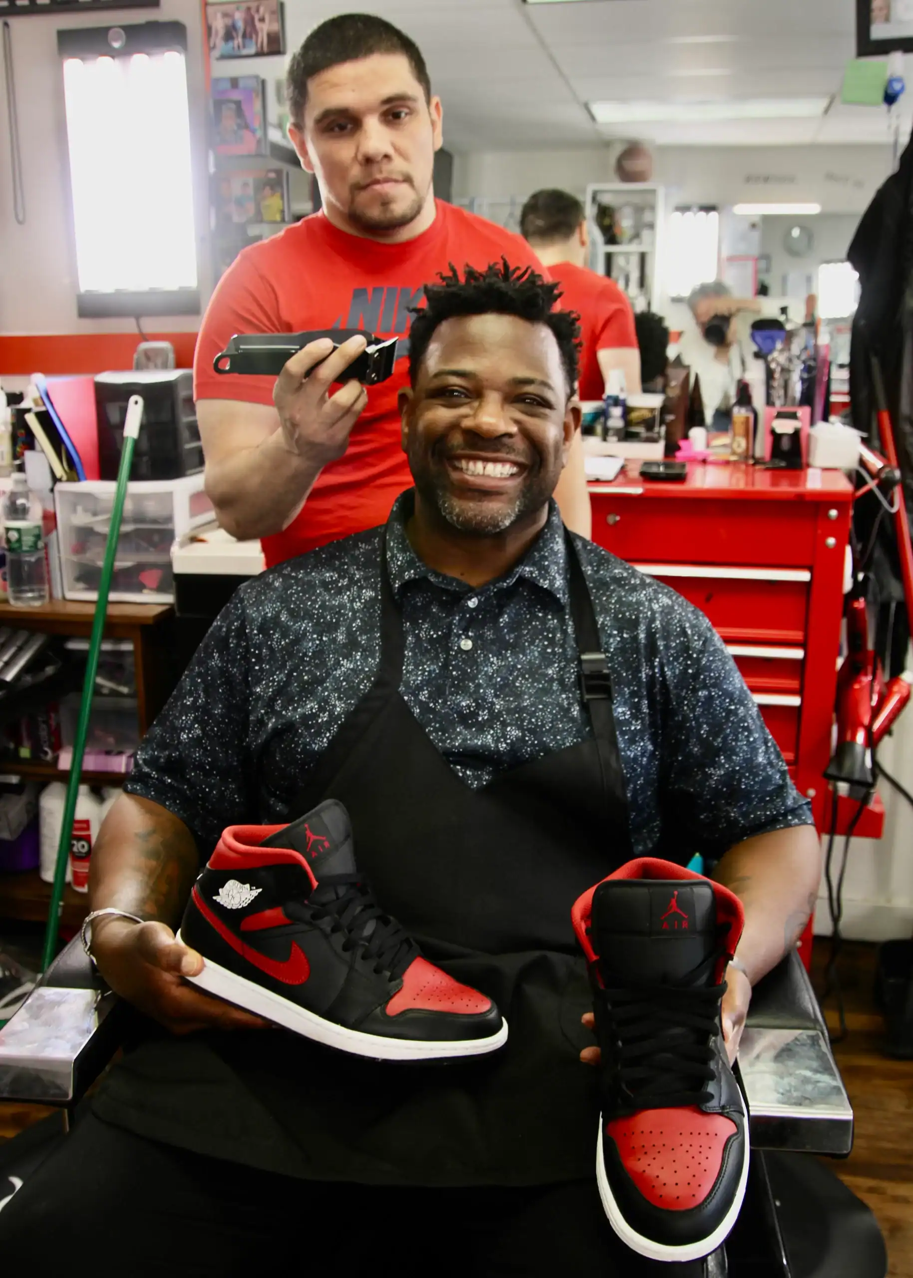 (Rick Miller/Olean Star) Church Without Walls Pastor Tyrone Hall shows off the types of sneakers Feet Heat Six is looking for this year as Hilario Maldonado trims a little off the top. This year’s sneaker distribution will be Aug. 24, at the church at 1803 W. State St.